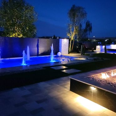 Nighttime backyard with blue-lit water fountains and a fire pit.