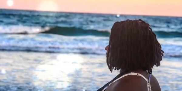 A woman with dreadlocks stands on a beach at sunset, gazing at the ocean.