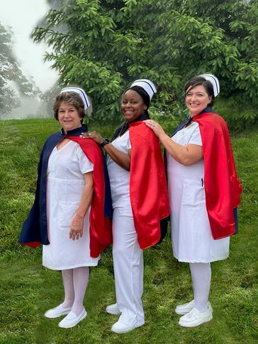 Three nurses in white uniforms wearing red and blue capes outdoors.