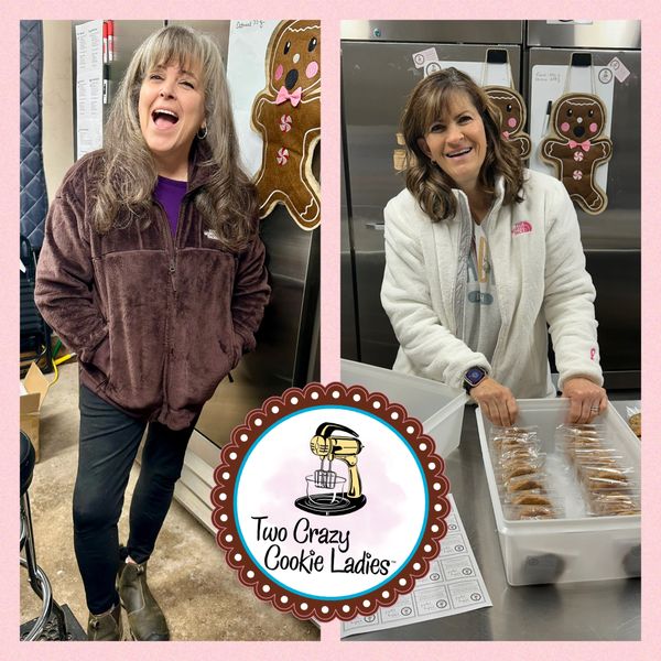 Two women smiling while preparing cookies in a kitchen with gingerbread decorations.