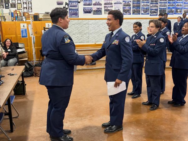 Two junior ROTC cadets shaking hands while others applaud in a classroom.
