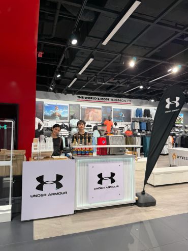 Two young people behind an Under Armour branded counter in a sports store.