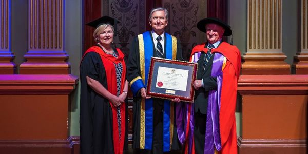 Three graduates in academic gowns posing with a framed certificate indoors.