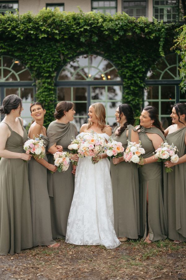 Bride and bridesmaids smiling, holding bouquets outside a greenery-covered building.