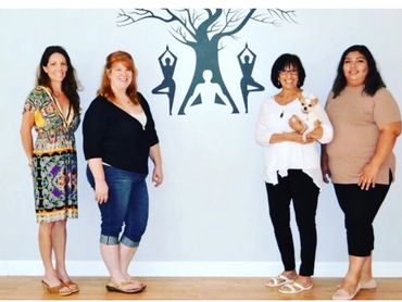 Four women standing in a yoga studio, one holding a small dog.