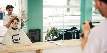 A barber cutting a young man's hair in a bright salon with a large mirror.