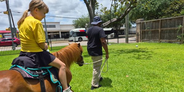 Child riding a pony led by an adult in a grassy area.