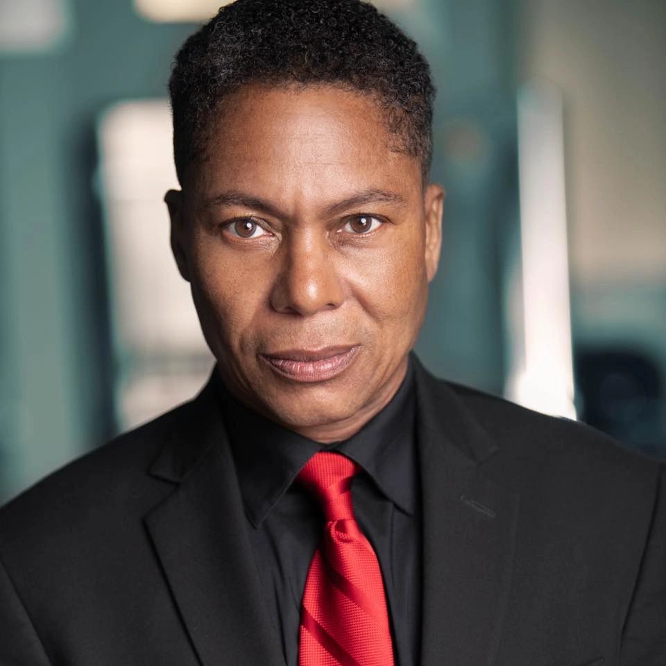 A confident man in a black suit and red tie looks directly at the camera.