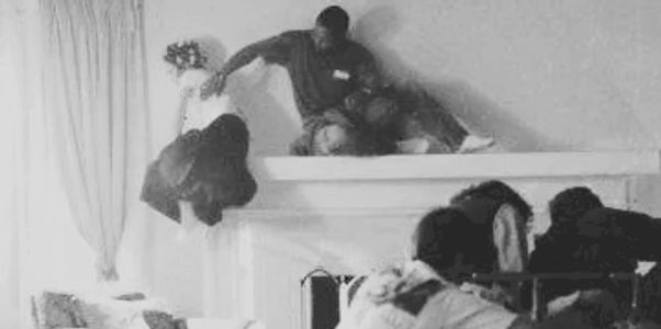 Crew filming a scene with actors on a fireplace mantel in a cozy living room.