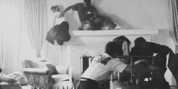 Crew filming a scene with actors on a fireplace mantel in a cozy living room.