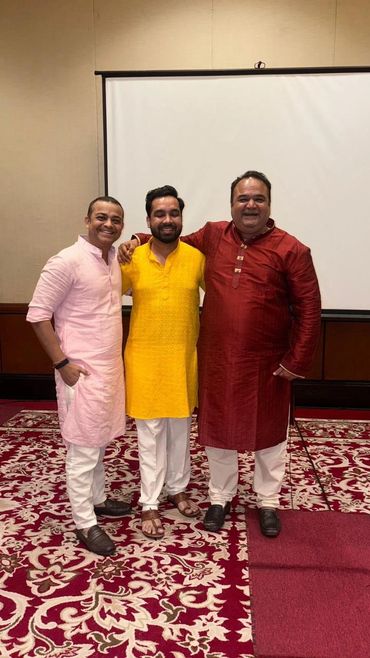 Three men in traditional Indian attire stand smiling together indoors.