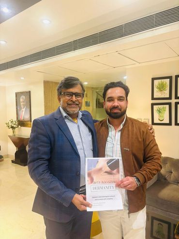 Two men smiling indoors, holding a dermatology magazine.