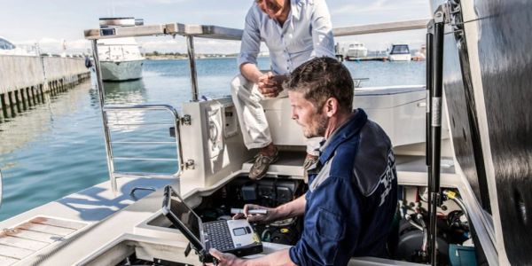 Two men working on a boat near a dock, one using a laptop.