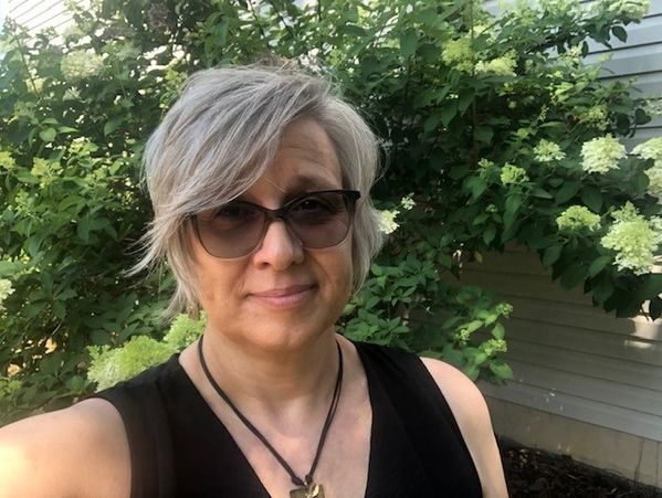 Occupational Therapy and Nervous System regulation therapist, a woman with gray hair and sunglasses smiling outdoors near green shrubs.