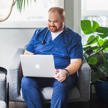Smiling male nurse in blue scrubs using a laptop while sitting in a chair.
