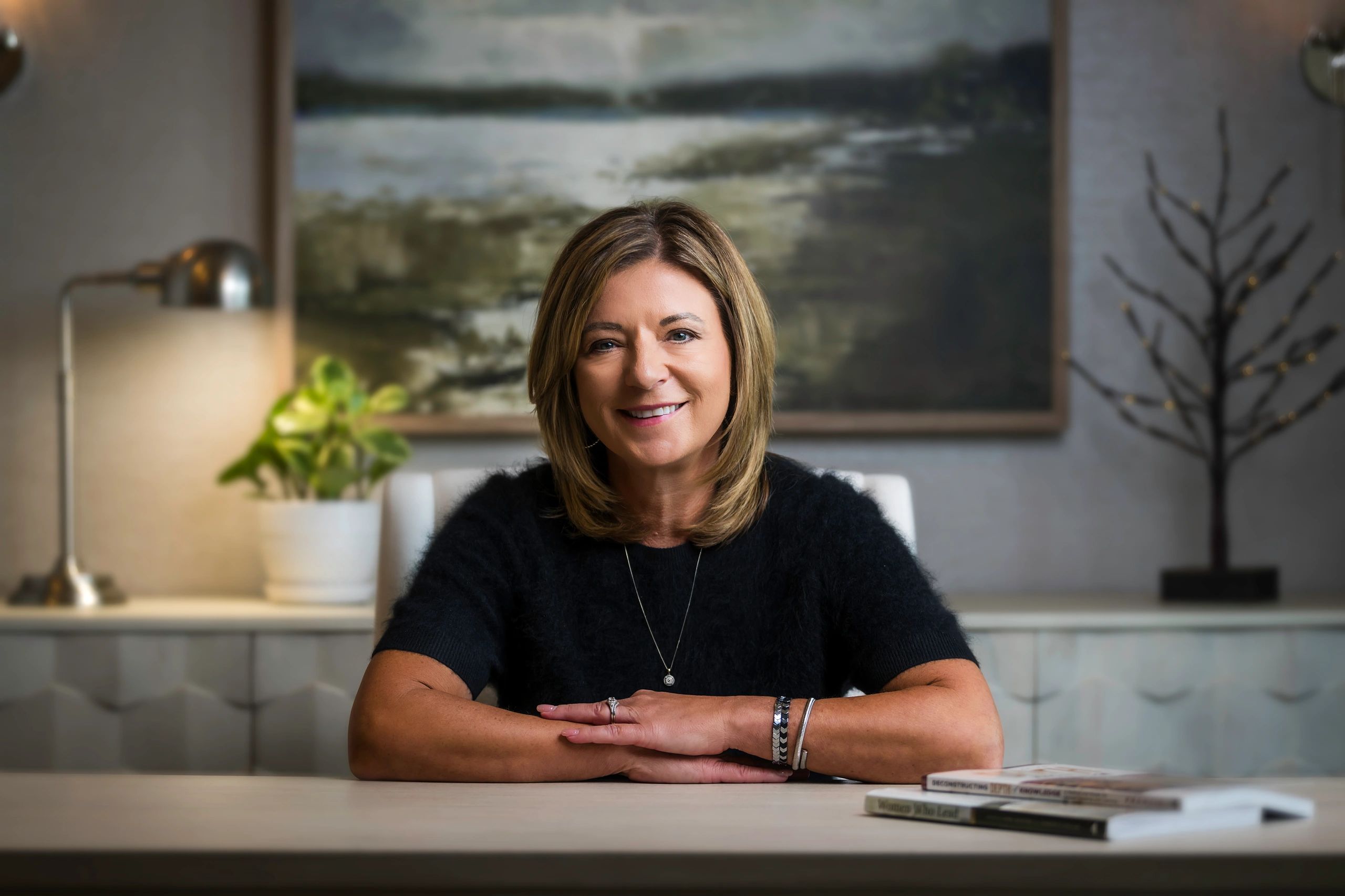 Smiling woman sitting at a desk with artwork and lamp in the background.