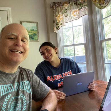 A man and a woman smiling at a wooden table with a laptop and sunny windows in the background.