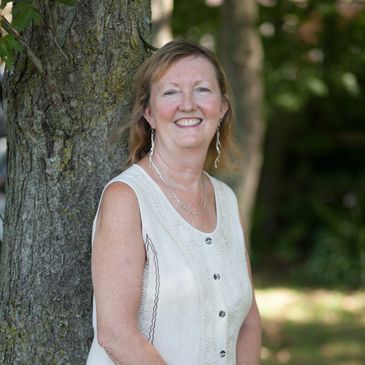 Smiling woman in a sleeveless dress standing by a tree outdoors.