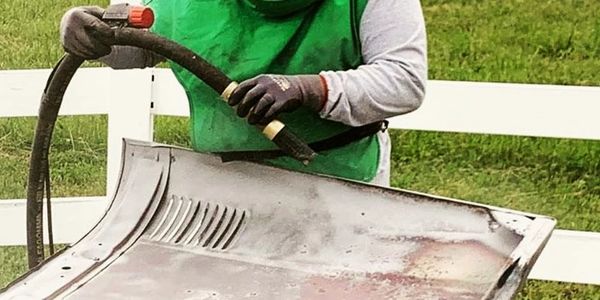 Person sandblasting a car hood outdoors with protective gear.