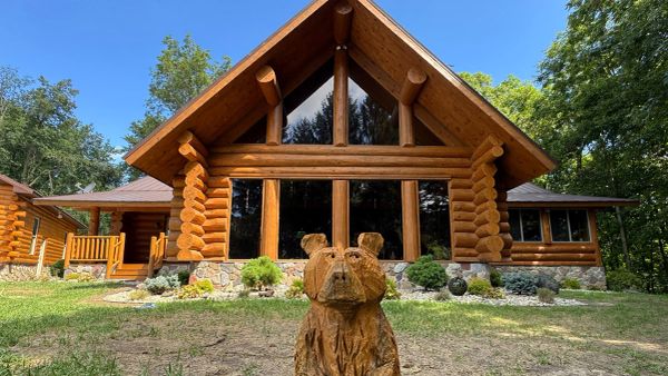 A wooden bear sculpture in front of a large log cabin surrounded by trees.