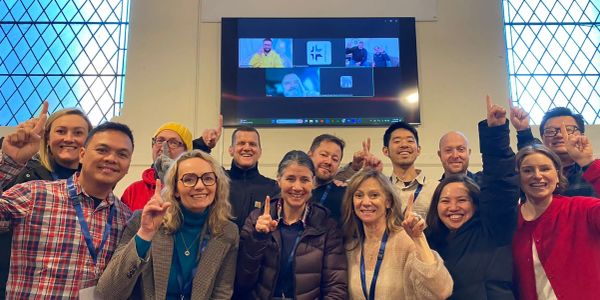 Group of people posing with fingers pointing up in front of a screen with virtual participants.