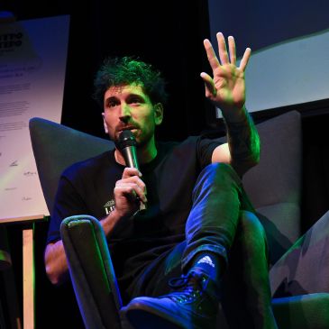 Man speaking into a microphone while seated on a stage.