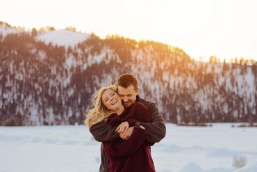 A couple embraces joyfully in a snowy mountain landscape at sunset.