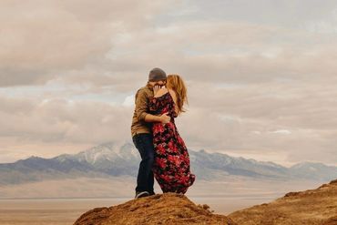 Couple embracing on a rocky hill with mountains in the background.