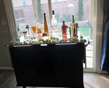 A home bar setup with various drinks and greenery on a table by a sliding glass door.