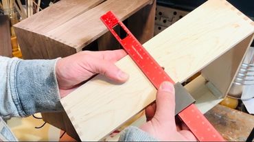 Person measuring a wooden box with a red ruler and square tool.