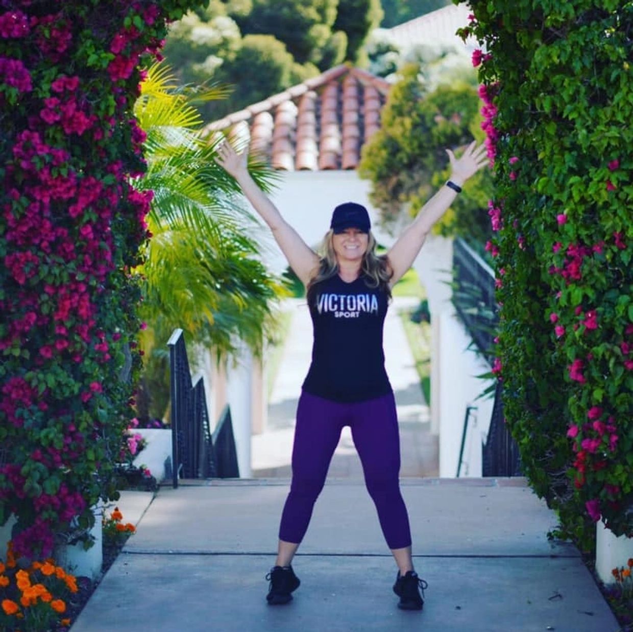 Woman in athletic wear smiling with arms raised outdoors between flower-covered pillars.