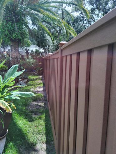 A brown wooden fence lines a lush garden with tropical plants and trees.