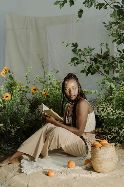 Black Woman Reading, sitting on blanket near a basket of oranges, with sunflowers in the background