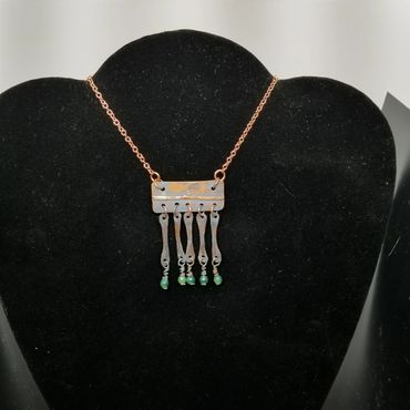 Unique metal necklace with dangling green beads on a black display.