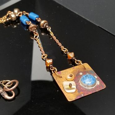 Artistic necklace with a square pendant featuring blue and amber accents on a gold-tone chain.