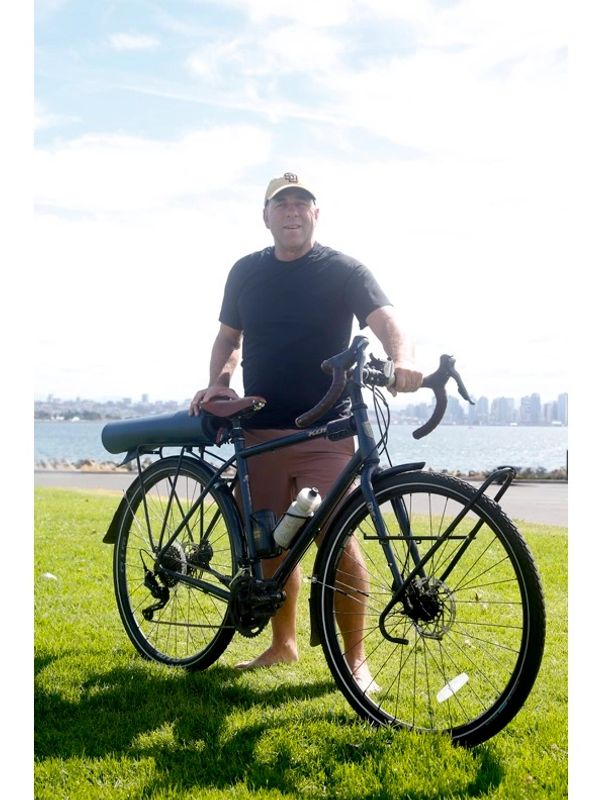 Man standing barefoot on grass with a black bicycle near the water.