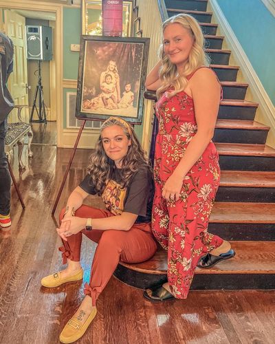 Two women pose by a staircase with a vintage portrait in the background.