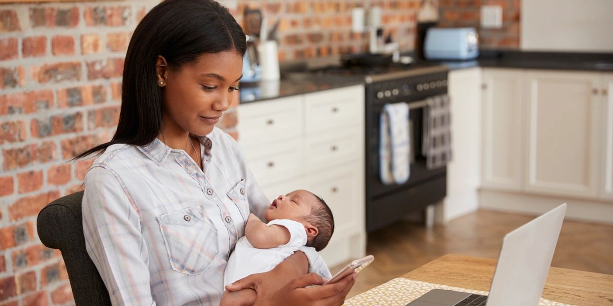 African American woman holding a baby while working on her cell phone and laptop in her home kitchen