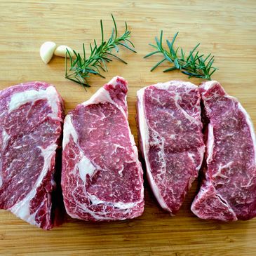 Four raw ribeye steaks with garlic and rosemary on a wooden board.