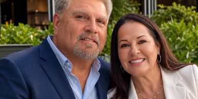 A smiling couple dressed formally, posing outdoors with greenery in the background.