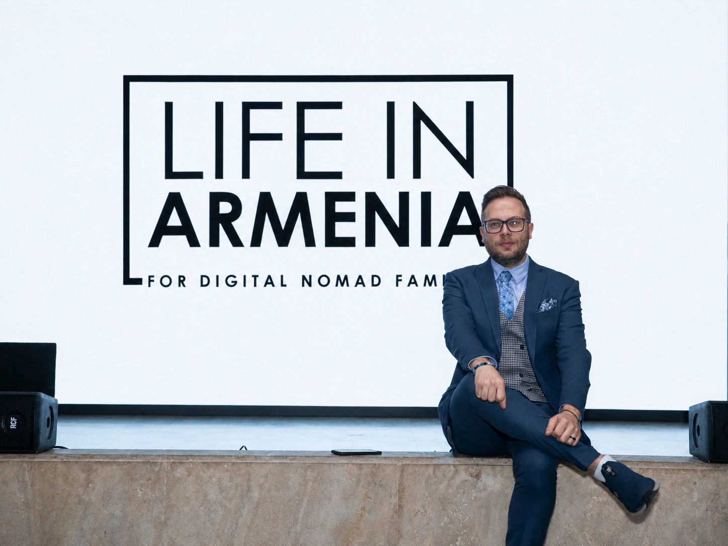 Man in suit sitting in front of a 'Life in Armenia' sign.