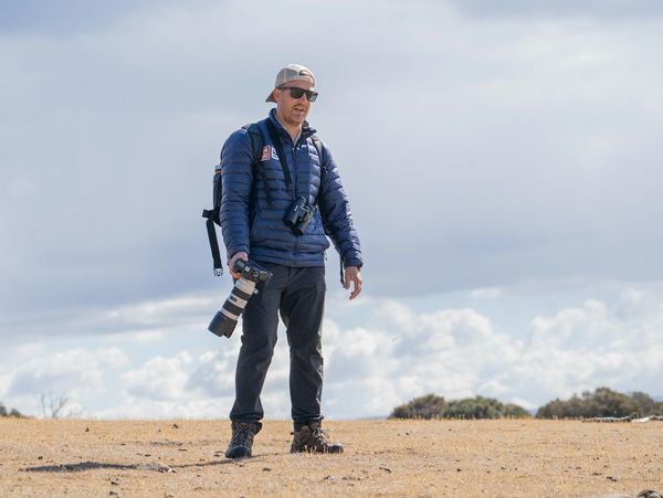 Man holding a camera in an open field under a cloudy sky.