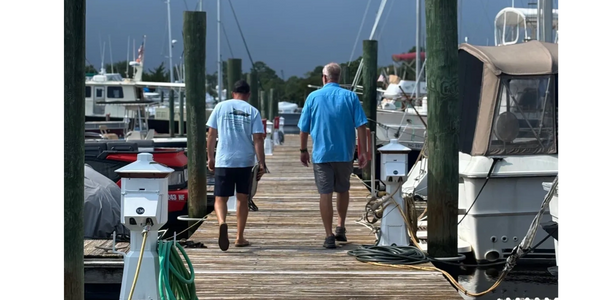 Two men walk down a wooden dock by moored boats under a cloudy sky.