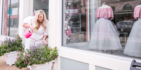 lady watering plants in front of pink and white dresses in a store front window