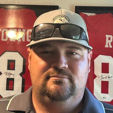 Man wearing a Pelican Bay cap and sunglasses with signed sports jerseys in the background.