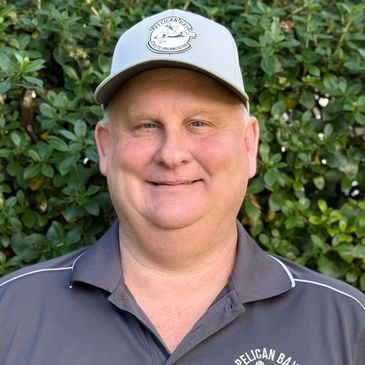 Smiling man in a grey polo and baseball cap outdoors against greenery.