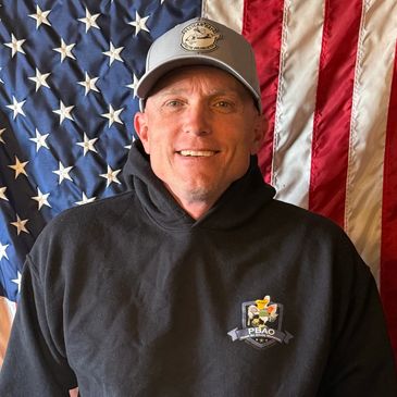 Smiling man in a black hoodie and cap in front of an American flag.