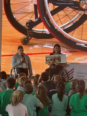 Patty holding a book and guest speaker with mic sitting in front of a group of kids