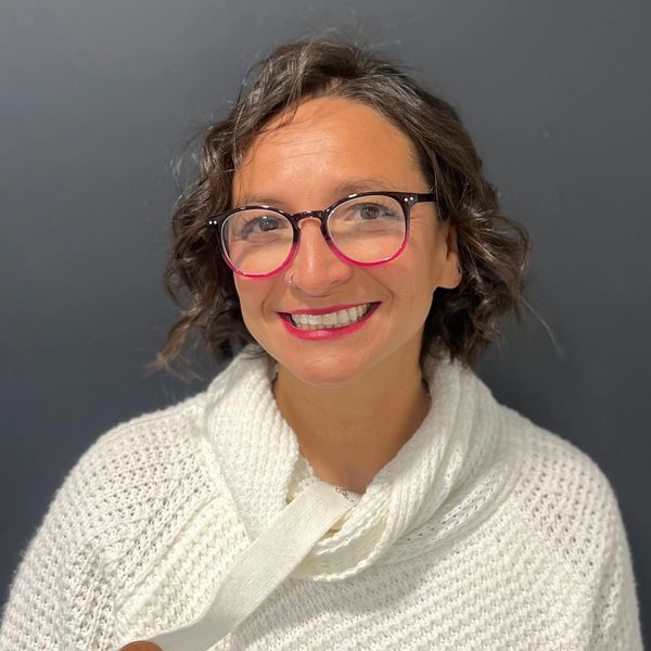Smiling woman with curly hair and colorful glasses wearing a cozy white sweater.