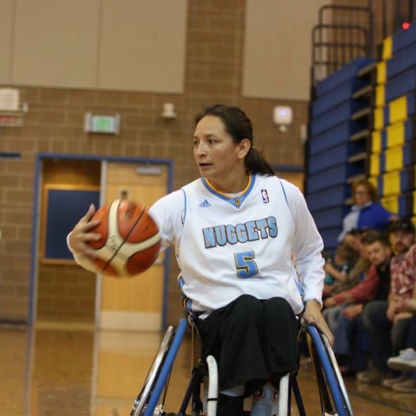 Patty sitting in a basketball wheelchair with a ball in hand wearing a white Nuggets jersey.